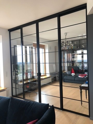 Przesuwne, szklane drzwi wewnętrzne z czarnymi, metalowymi szprosami, dzielące przestrzeń salonu i gabinetu, widoczne odbicie wnętrza w szkle.
