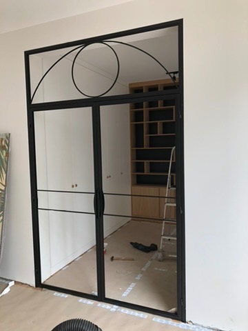 Dwuskrzydłowe drzwi szklane z czarną metalową ramą i dekoracyjnym łukiem u góry, widoczne ślady prac wykończeniowych na podłodze.