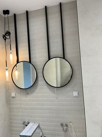 Dwa okrągłe lustra z czarną ramą zawieszone na długich, czarnych metalowych prętach, zamontowane na ścianie z szarych płytek imitujących cegłę, widoczne instalacje hydrauliczne i elektryczne...