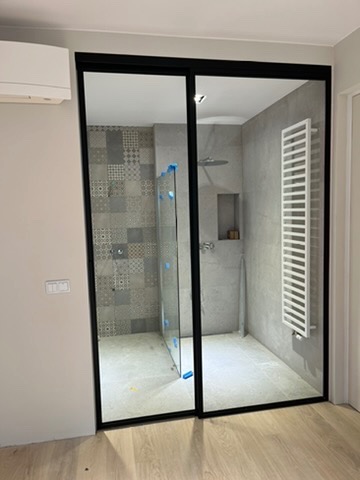 Nowoczesna kabina prysznicowa ze szklanymi drzwiami przesuwnymi w czarnej ramie, zrealizowana w Starych Babicach. Widoczna mozaika na ścianie i grzejnik drabinkowy.