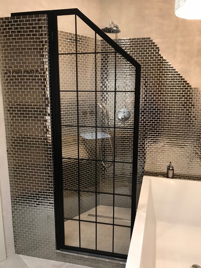 Kabina prysznicowa z czarną, metalową ramą i szklanymi panelami, otoczona ścianami wyłożonymi błyszczącą mozaiką w odcieniach szarości, widoczny odpływ liniowy w podłodze i fragment białej wanny.