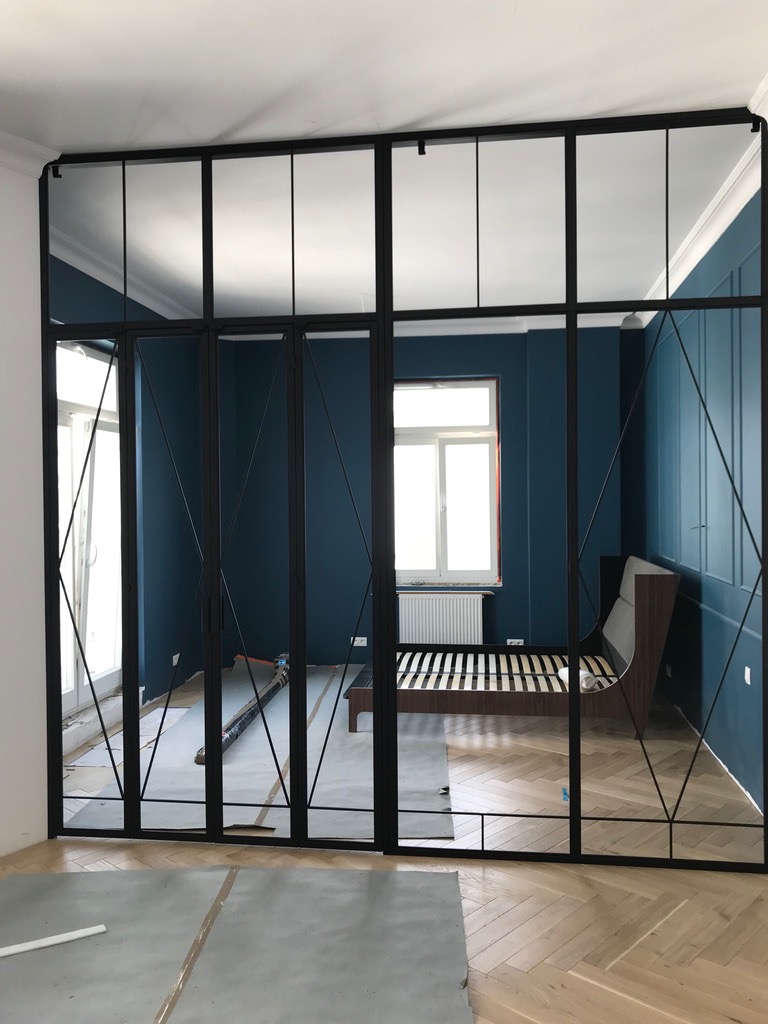 Szklane drzwi z czarnymi metalowymi ramami oddzielające sypialnię od reszty mieszkania, widoczne zabezpieczenia podłogi podczas montażu.