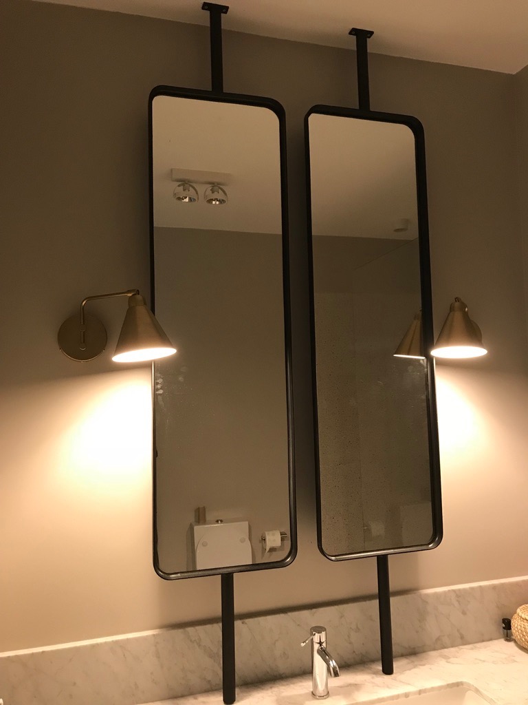 Dwa wysokie, prostokątne lustra z czarną ramą, zamontowane na metalowych wspornikach, oświetlone złotymi kinkietami, odbijające wnętrze łazienki z marmurowym blatem i nowoczesną armaturą.
