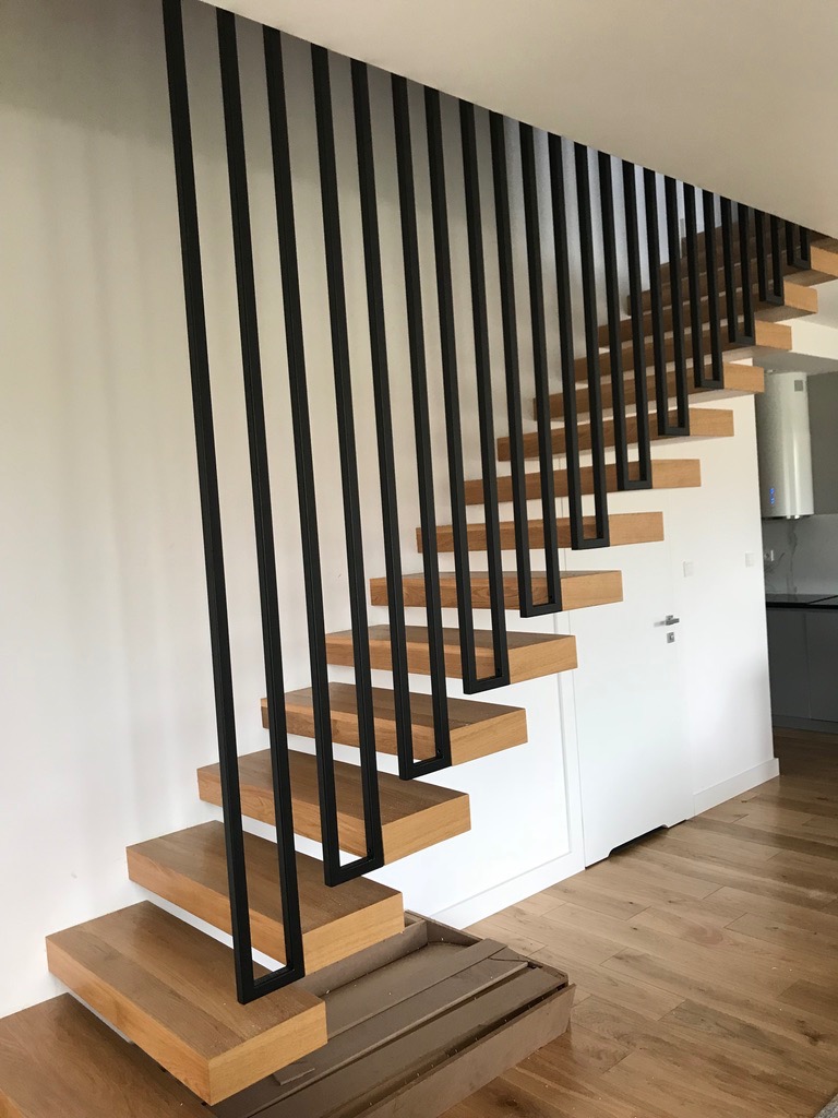 Nowoczesne schody drewniane z czarną, minimalistyczną balustradą metalową, stopnie bez podstopnic, widoczne fragmenty podłogi i ściany w jasnych kolorach, w tle drzwi i fragment kuchni.