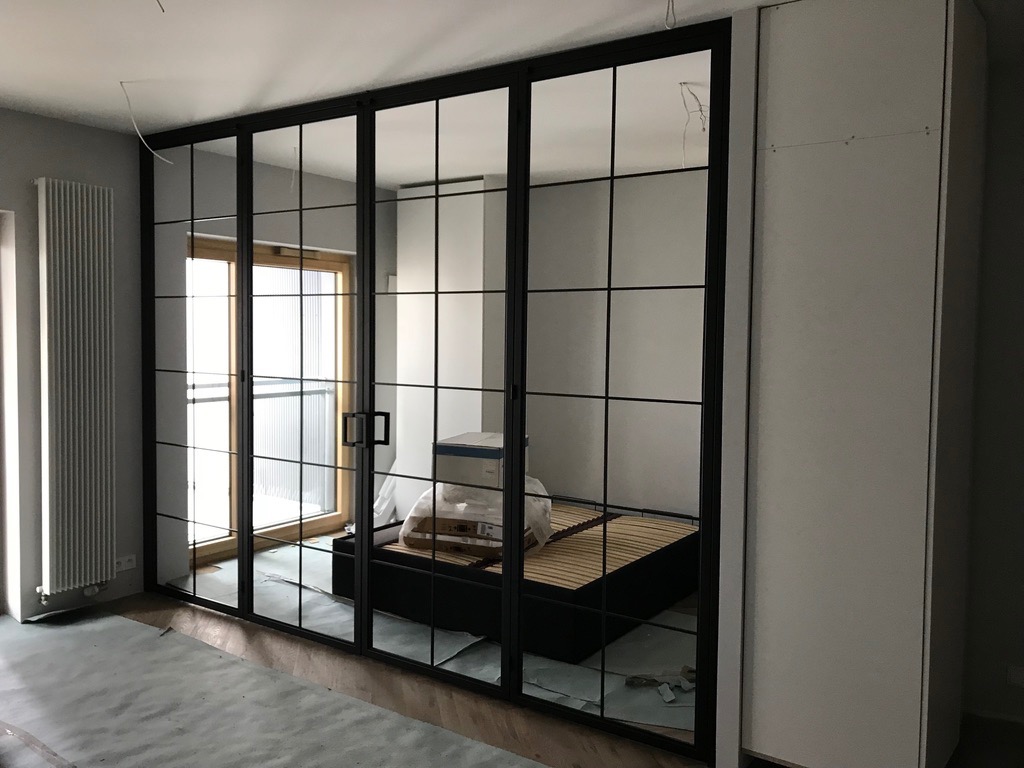 Nowoczesne drzwi szklane w czarnej metalowej ramie z widoczną sypialnią w tle, częściowo zakryte folią ochronną na podłodze.