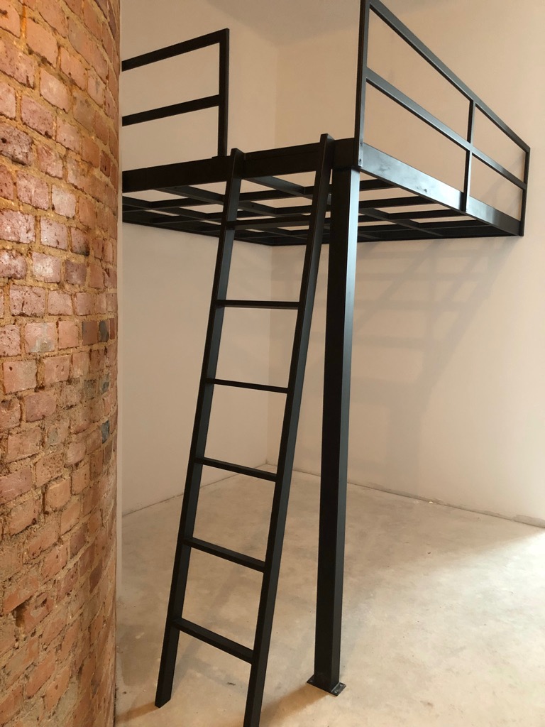Czarna metalowa konstrukcja antresoli z drabinką, umieszczona w pokoju z ceglaną ścianą, widok z dołu.