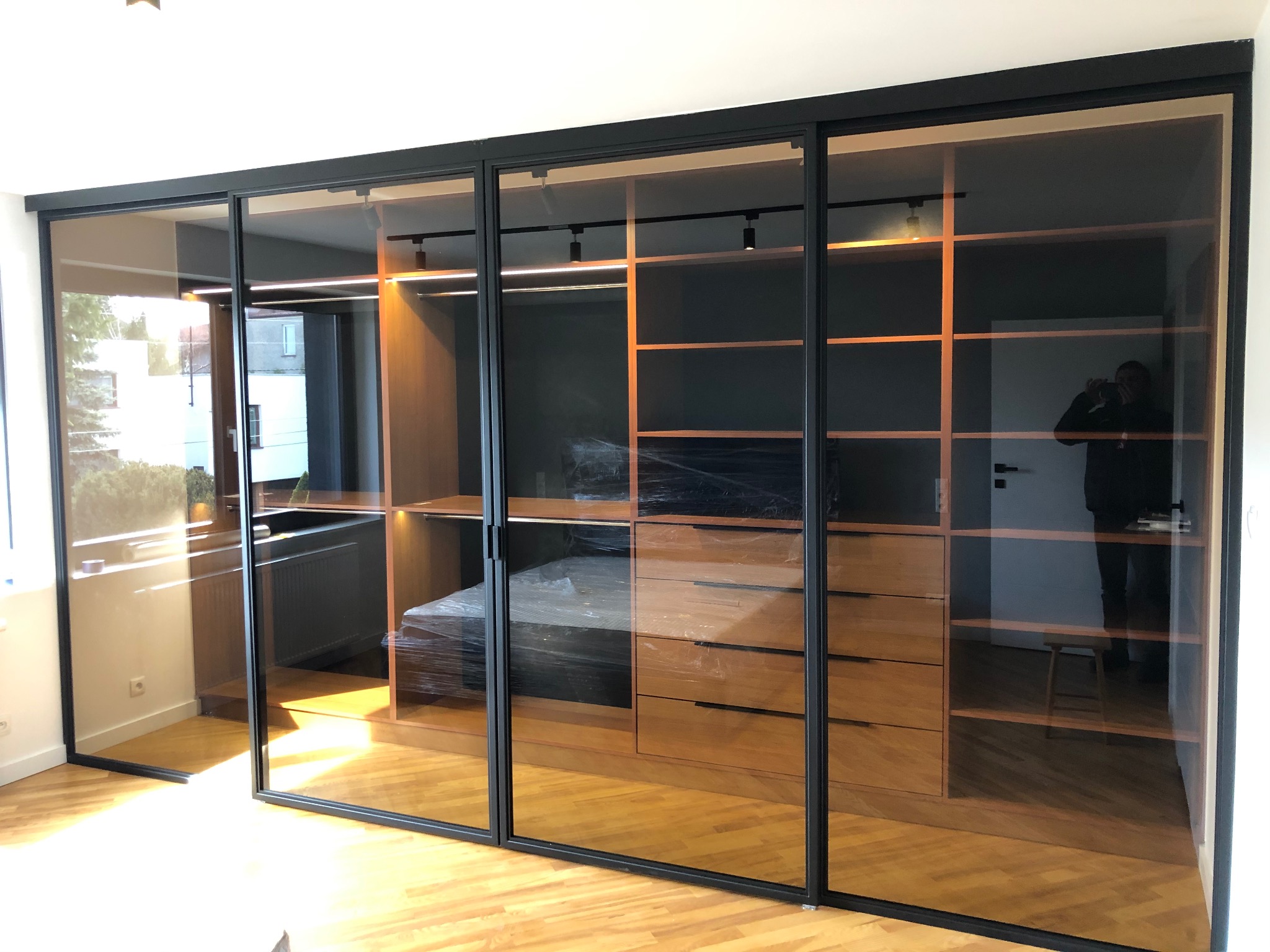 Szklane drzwi przesuwne z czarnymi metalowymi ramami oddzielające garderobę z drewnianymi półkami i komodą, widok z parkietu w jodełkę.