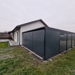 Biuro Projektów DOMARG Radosław Grzanka - Nowoczesna, grafitowa pergola metalowa, przylegająca do białego domu z czarnym dachem. Widok z boku, trawnik, kostka brukowa i zarys jesiennego krajobrazu w tle.