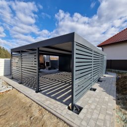 Biuro Projektów DOMARG Radosław Grzanka - Nowoczesna, grafitowa pergola metalowa z poziomymi lamelami, częściowo zabudowana, na tle błękitnego nieba z chmurami. Widoczne kostki brukowe i przygotowany teren.