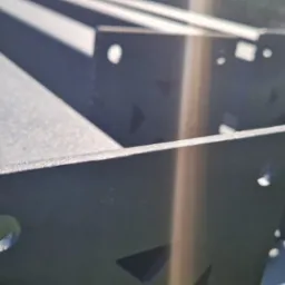 Czarne, metalowe elementy konstrukcyjne z otworami, prawdopodobnie do pergoli, leżące na trawie w słoneczny dzień. Widoczne detale wykończenia.