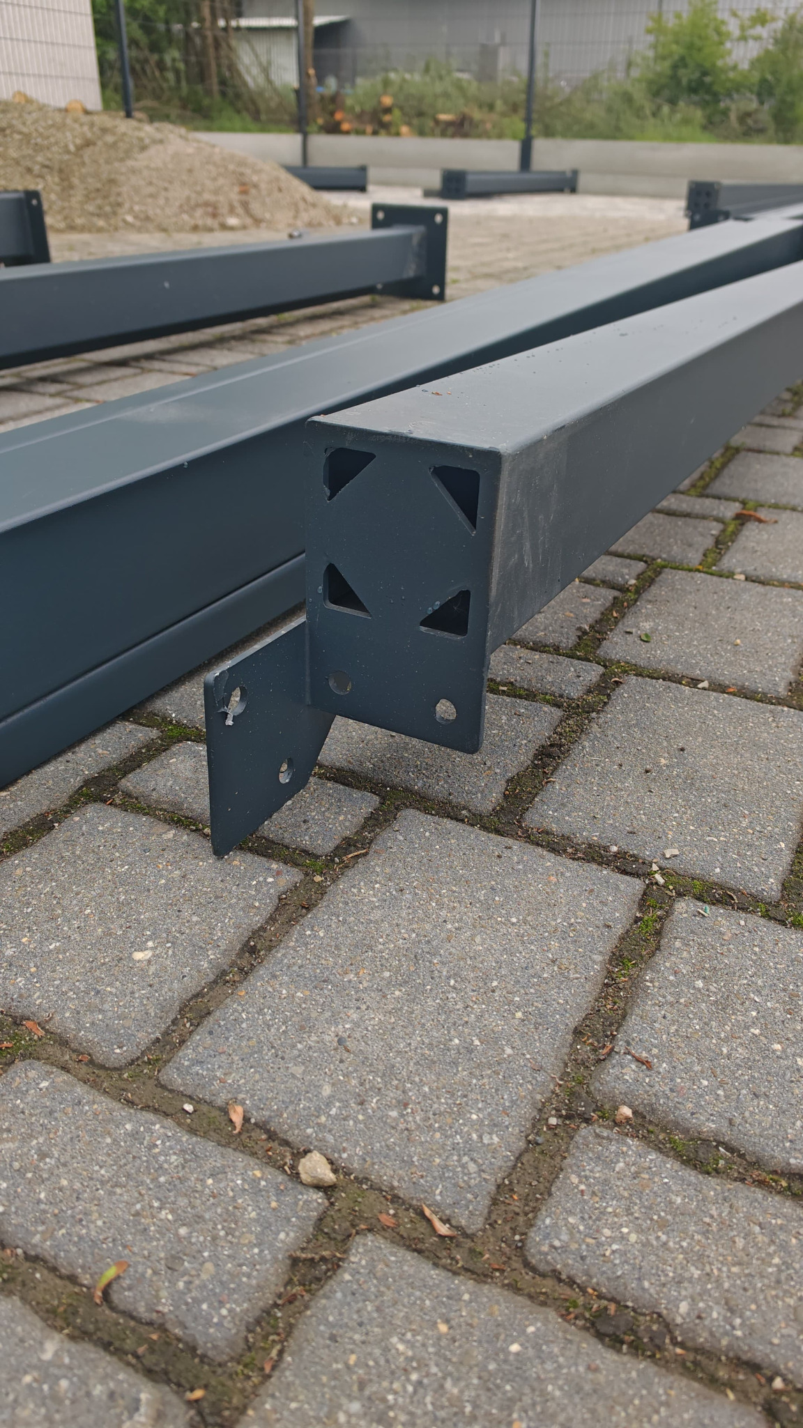 Szare, metalowe profile konstrukcyjne z mocowaniem, leżące na szarej kostce brukowej. Widoczne otwory montażowe. W tle ziemia i zieleń.