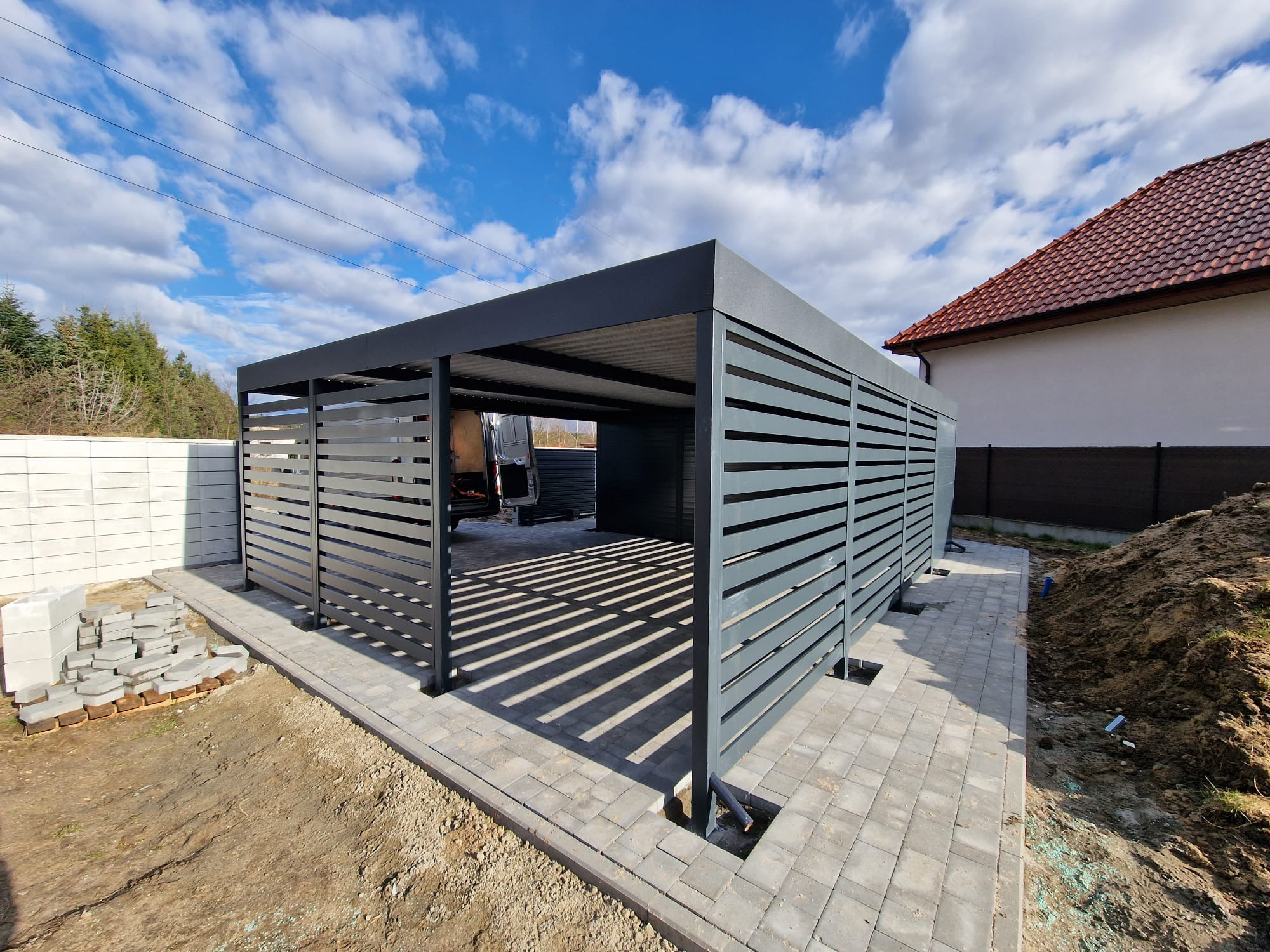 Nowoczesna, grafitowa pergola metalowa z poziomymi lamelami, częściowo zabudowana, na tle błękitnego nieba z chmurami. Widoczne kostki brukowe i przygotowany teren.