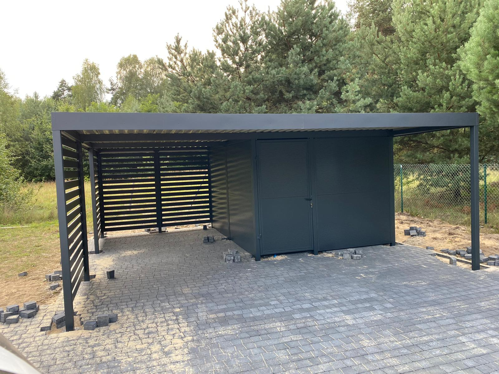 Nowoczesna, grafitowa pergola metalowa z zamykaną częścią gospodarczą i ażurowymi panelami bocznymi, wykończona kostką brukową na tle zieleni.