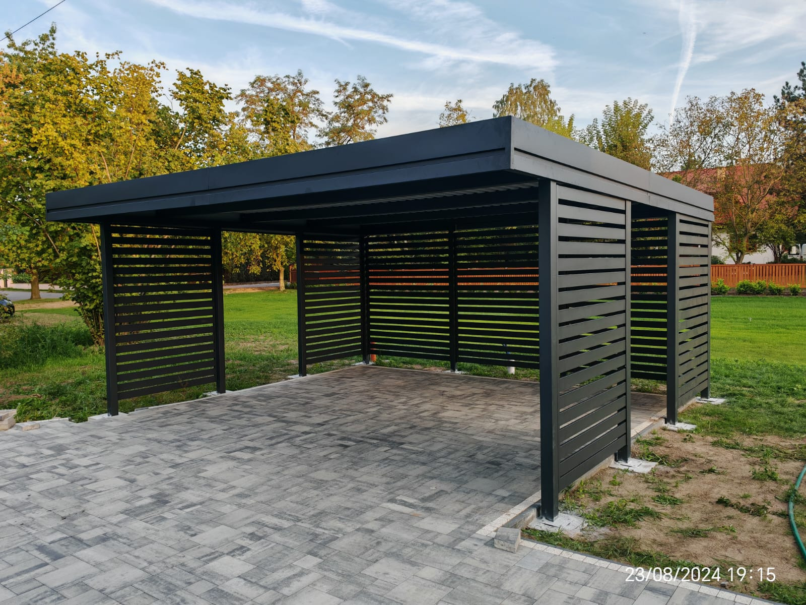 Nowoczesna, grafitowa pergola metalowa z poziomymi lamelami, na tle zielonego trawnika i kostki brukowej. Widoczne detale konstrukcji i staranne wykończenie.