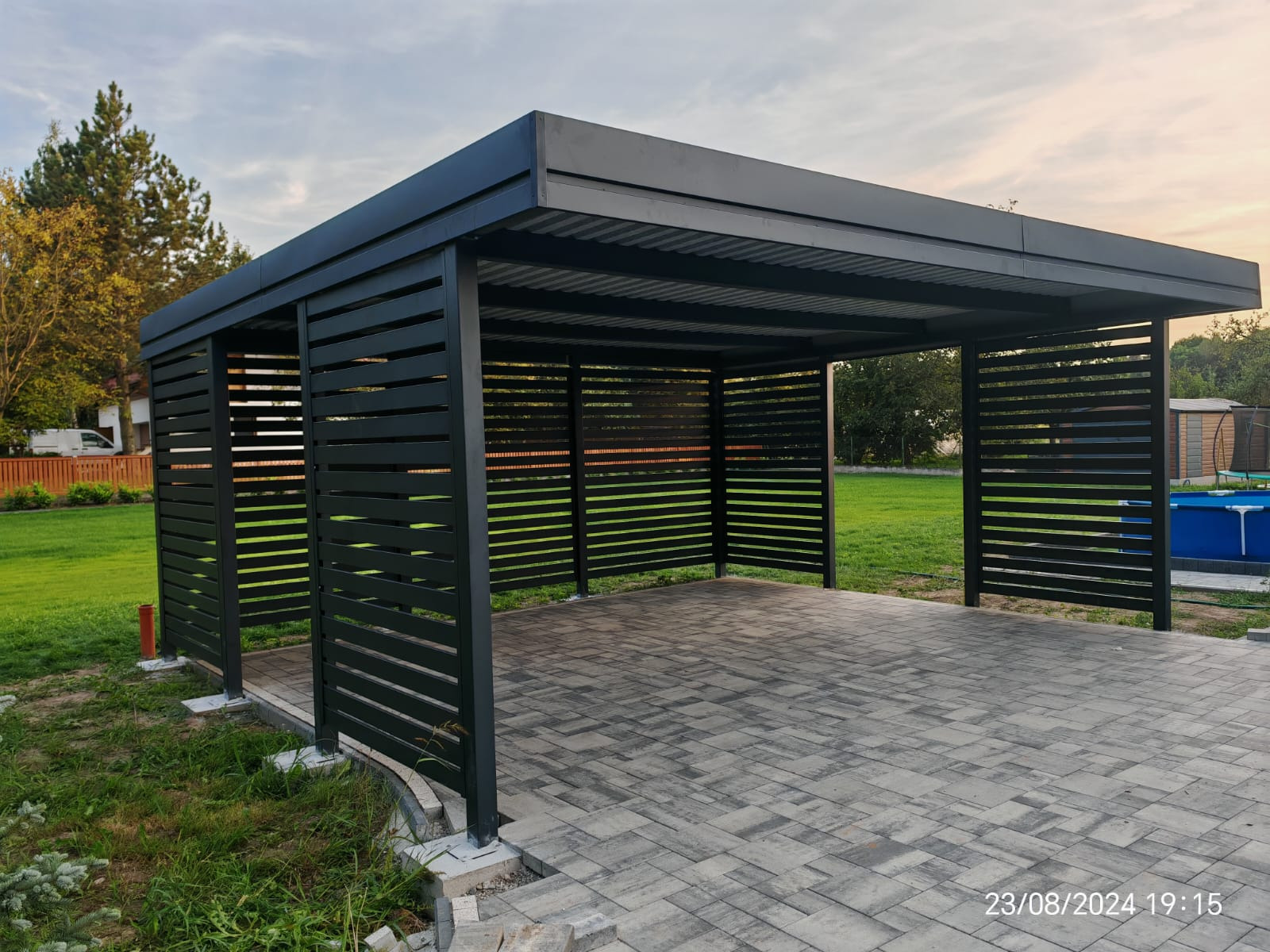 Nowoczesna, grafitowa pergola metalowa z poziomymi listwami, stojąca na kostce brukowej. W tle trawnik i drzewa, widoczne elementy ogrodu.