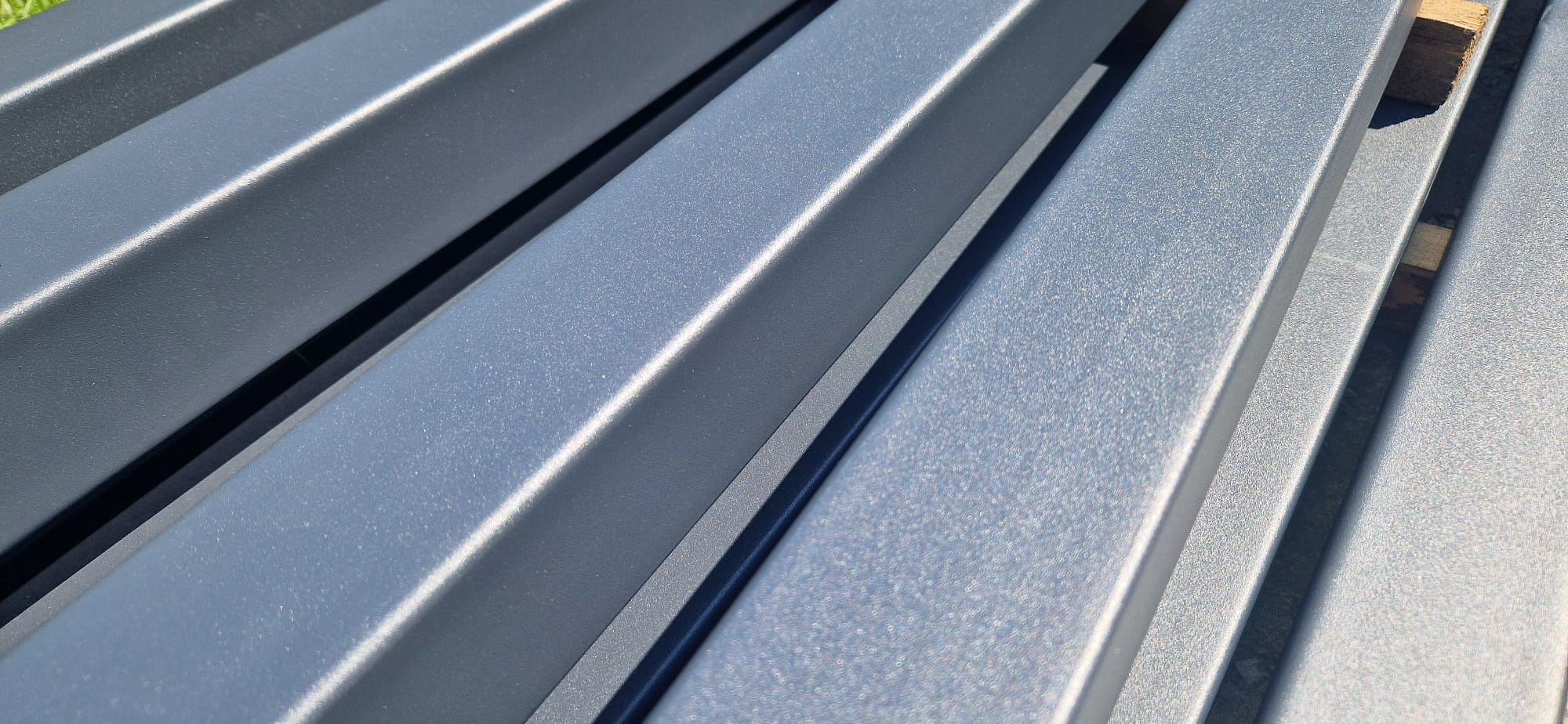 Szare, metalowe profile konstrukcyjne z widoczną fakturą, ułożone równolegle. Widoczne cienie i ostre światło słoneczne podkreślają geometrię elementów.