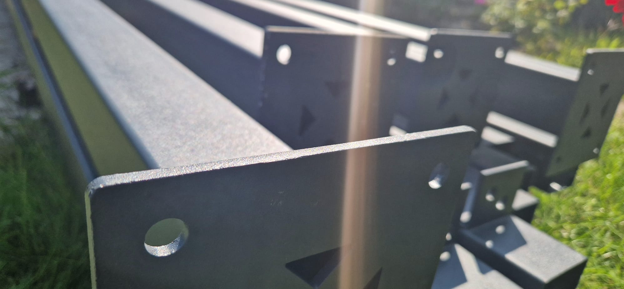 Czarne, metalowe elementy konstrukcyjne z otworami, prawdopodobnie do pergoli, leżące na trawie w słoneczny dzień. Widoczne detale wykończenia.