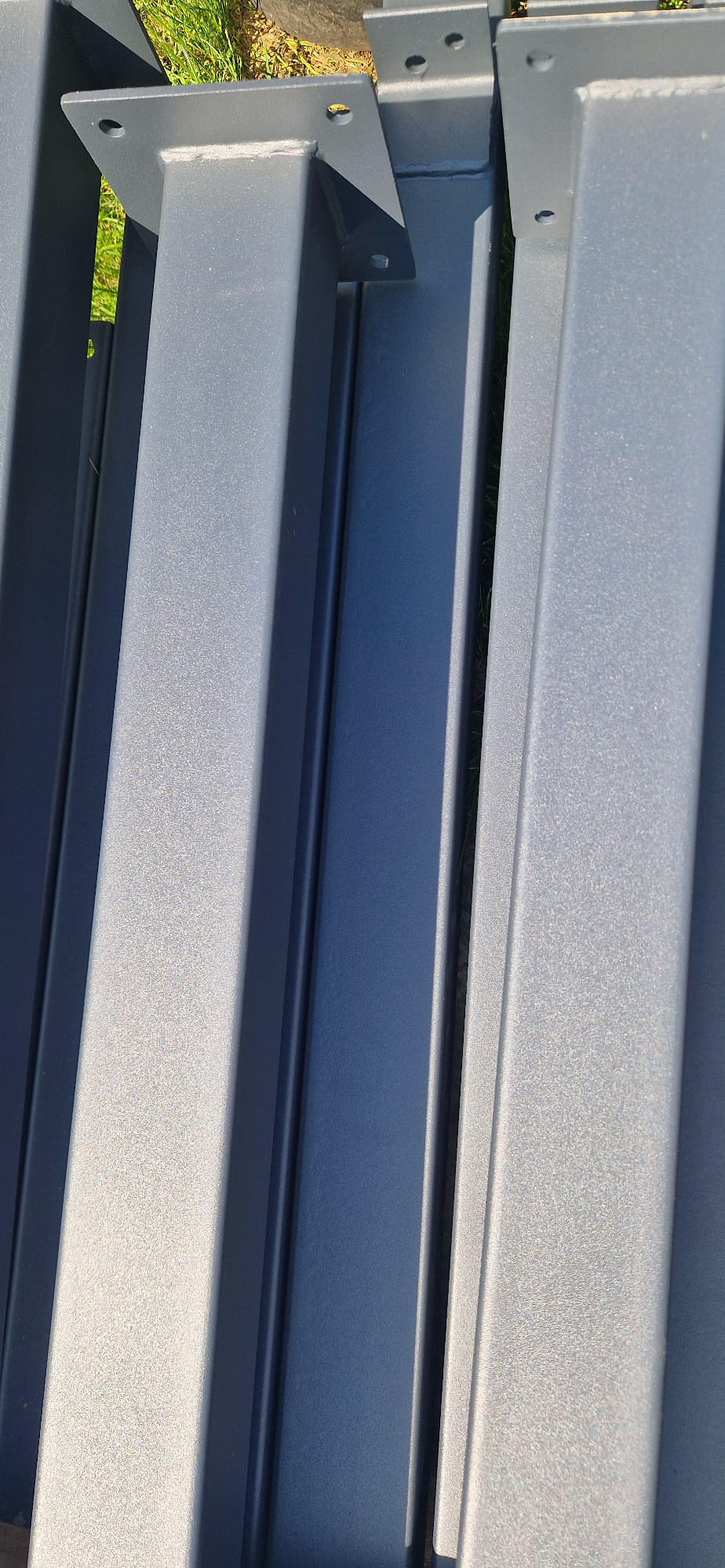 Metalowe słupy w kolorze grafitowym z podstawami montażowymi, leżące na trawie. Elementy konstrukcyjne do budowy pergoli lub wiaty.