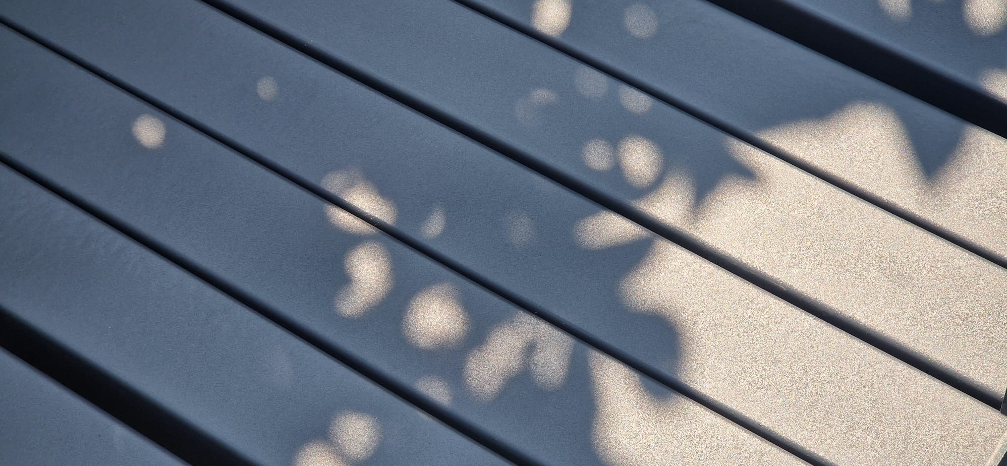 Metalowa konstrukcja z cieniem liści, prawdopodobnie element pergoli lub zabudowy balkonu. Widoczne regularne, równoległe profile.