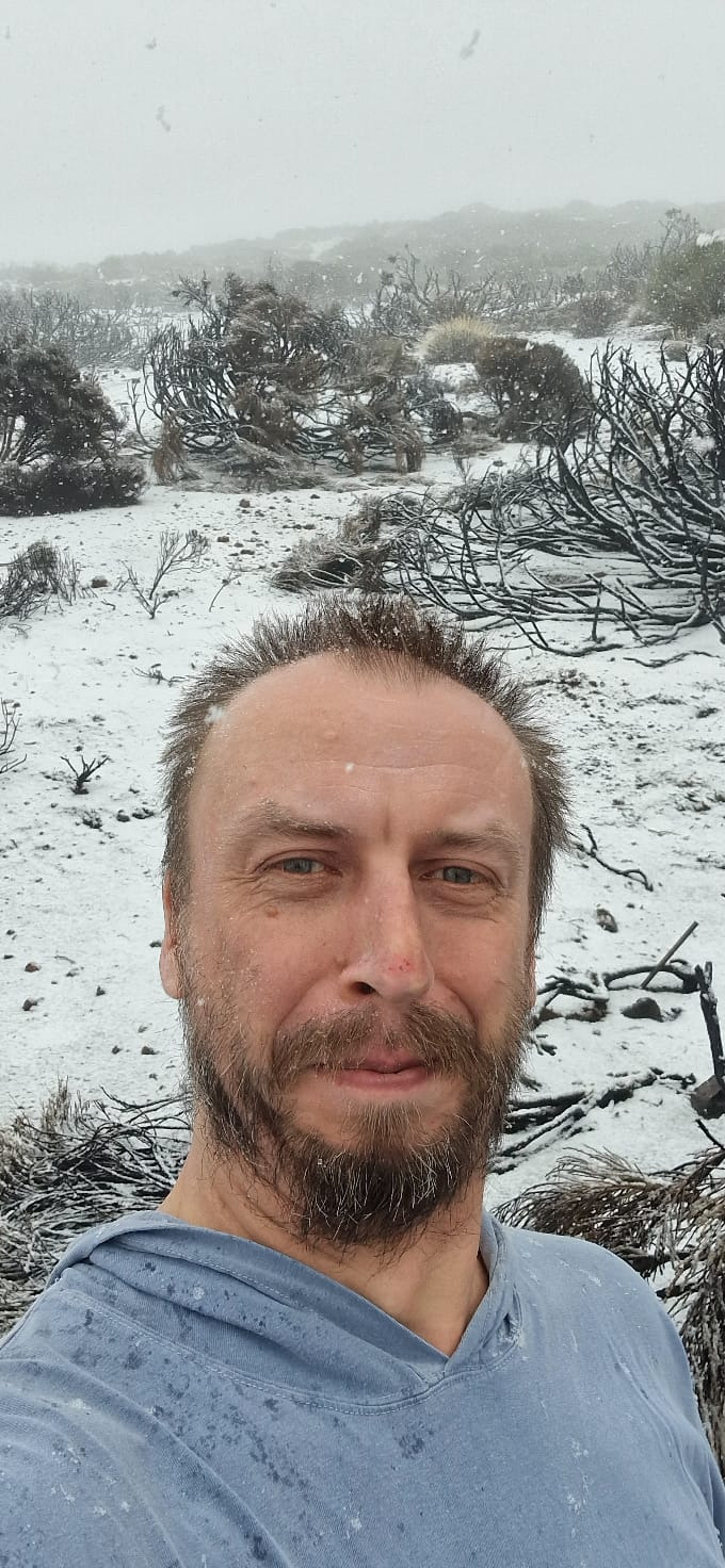 Portret mężczyzny z brodą na tle zimowego krajobrazu. W tle krzewy przysypane śniegiem, w powietrzu opad śniegu. Ujęcie z bliska, widoczna twarz i część ramion.