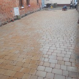 Brukarstwo Karol Brzozowski - Dziedziniec wyłożony brązową kostką brukową, widoczni pracownicy podczas prac wykończeniowych, budynki z cegły i otynkowane w tle.