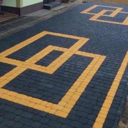 Brukarstwo Karol Brzozowski - Ułożona nawierzchnia z ciemnoszarej kostki brukowej z geometrycznym wzorem z jasnej kostki, widoczny budynek gospodarczy i pracownicy w tle.