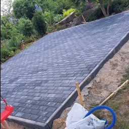 Brukarstwo Karol Brzozowski - Powierzchnia wyłożona szarą kostką brukową na nierównym terenie, widoczny fragment huśtawki i niebieski wąż, w tle zieleń drzew i krzewów.