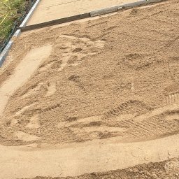 Brukarstwo Karol Brzozowski - Przygotowany teren pod ścieżkę z kostki brukowej, wyrównany piasek z widocznymi śladami opon, kostka ułożona częściowo w oddali, osoba w pomarańczowych spodniach w tle.