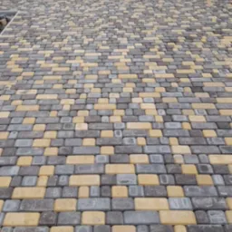 Ułożona nawierzchnia z kostki brukowej w odcieniach szarości i żółci, prowadząca do domu z czerwonymi kwiatami w donicach, widoczne złączenia kostek.