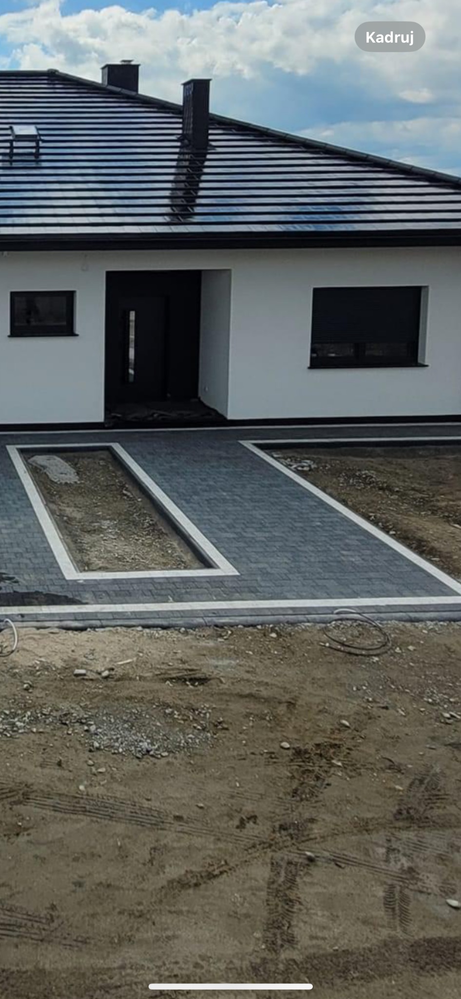 Nowoczesny dom jednorodzinny z szarą kostką brukową tworzącą chodnik i wjazdy, obrzeża wykończone jasnym kamieniem, teren wokół jeszcze niezagospodarowany.