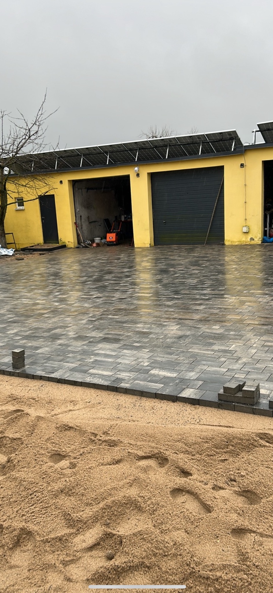 Układanie szarej kostki brukowej na piasku przed żółtym budynkiem z panelami słonecznymi, widoczna zagęszczarka w otwartym garażu.