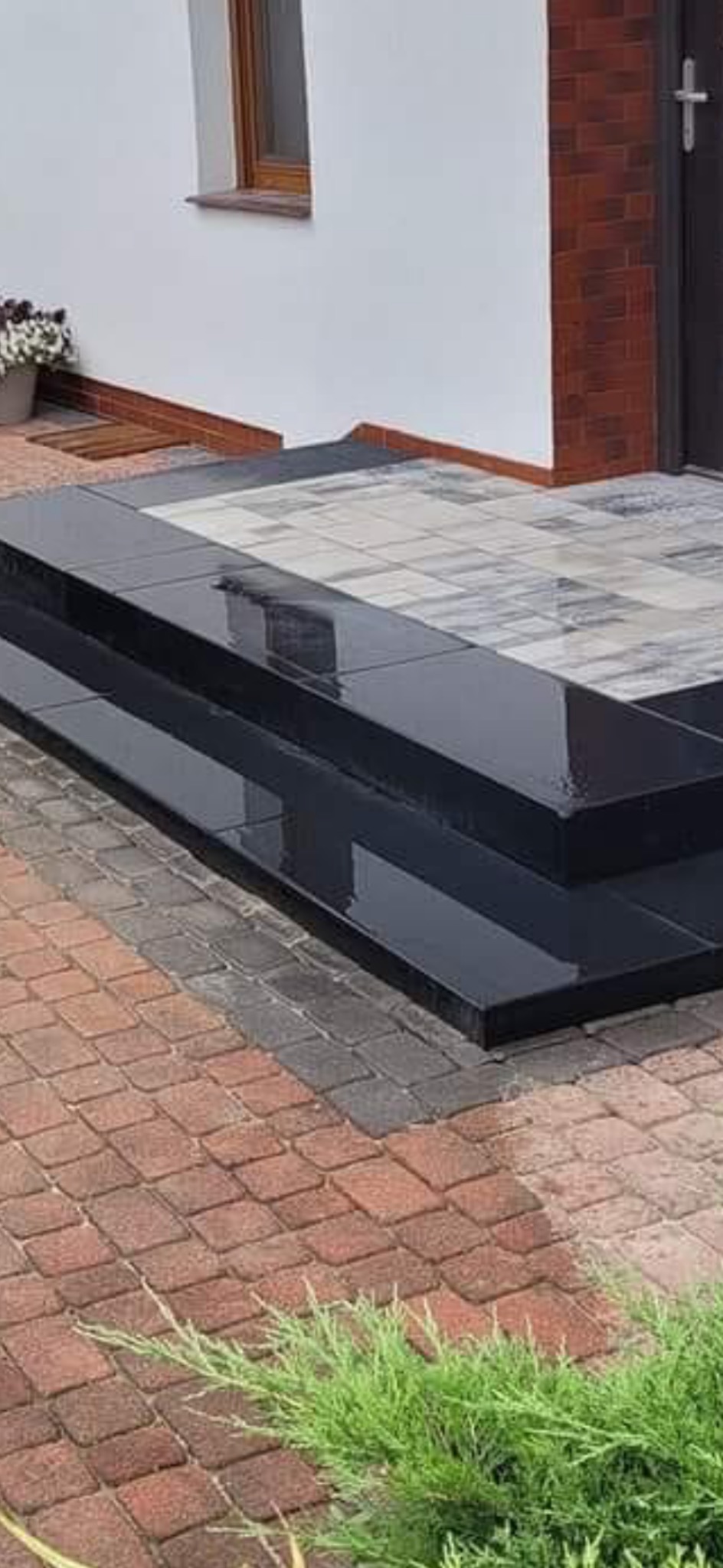 Dwustopniowe, czarne schody z polerowanego kamienia prowadzą do domu z ceglanym narożnikiem i kostką brukową w odcieniach czerwieni i szarości.