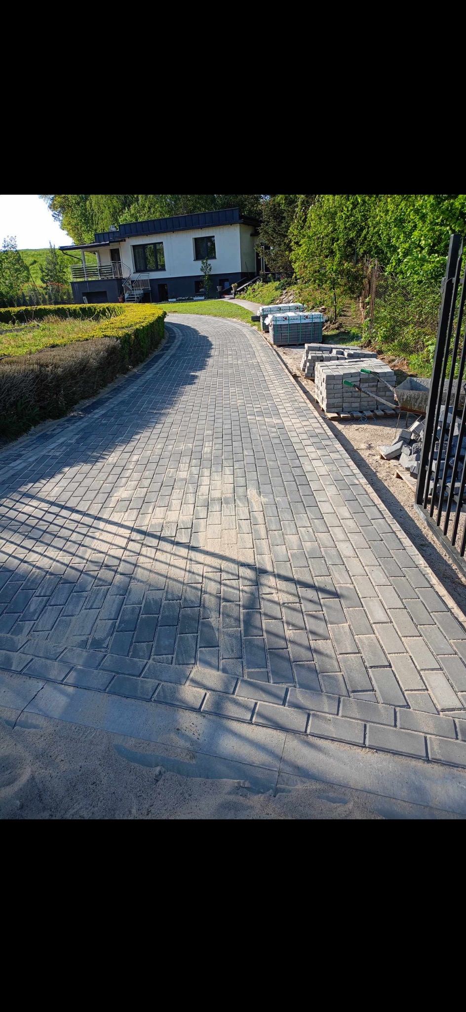 Ukończony podjazd z szarej kostki brukowej prowadzący do domu jednorodzinnego, z paletami kostki obok, widoczne cienie ogrodzenia.