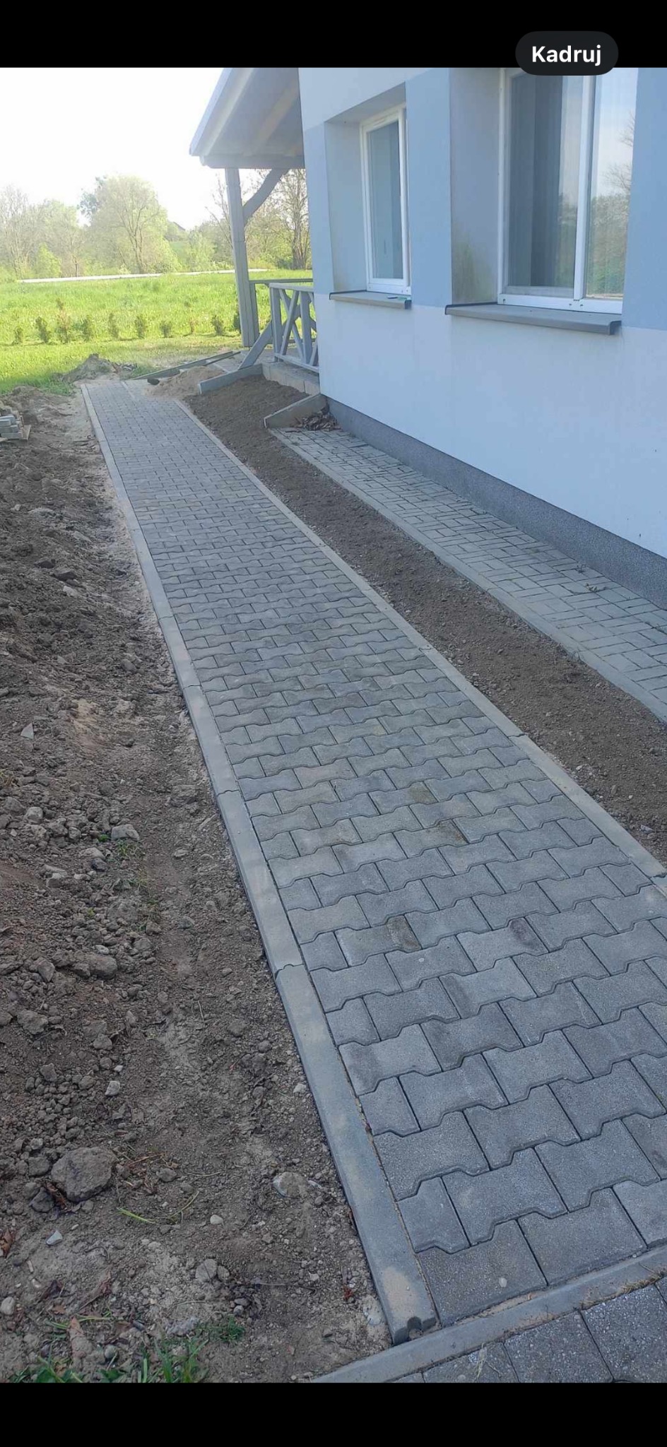 Nowa, szara, betonowa ścieżka z kostki brukowej prowadząca wzdłuż jasnoniebieskiego domu z białymi oknami, otoczona ziemią i zielenią.