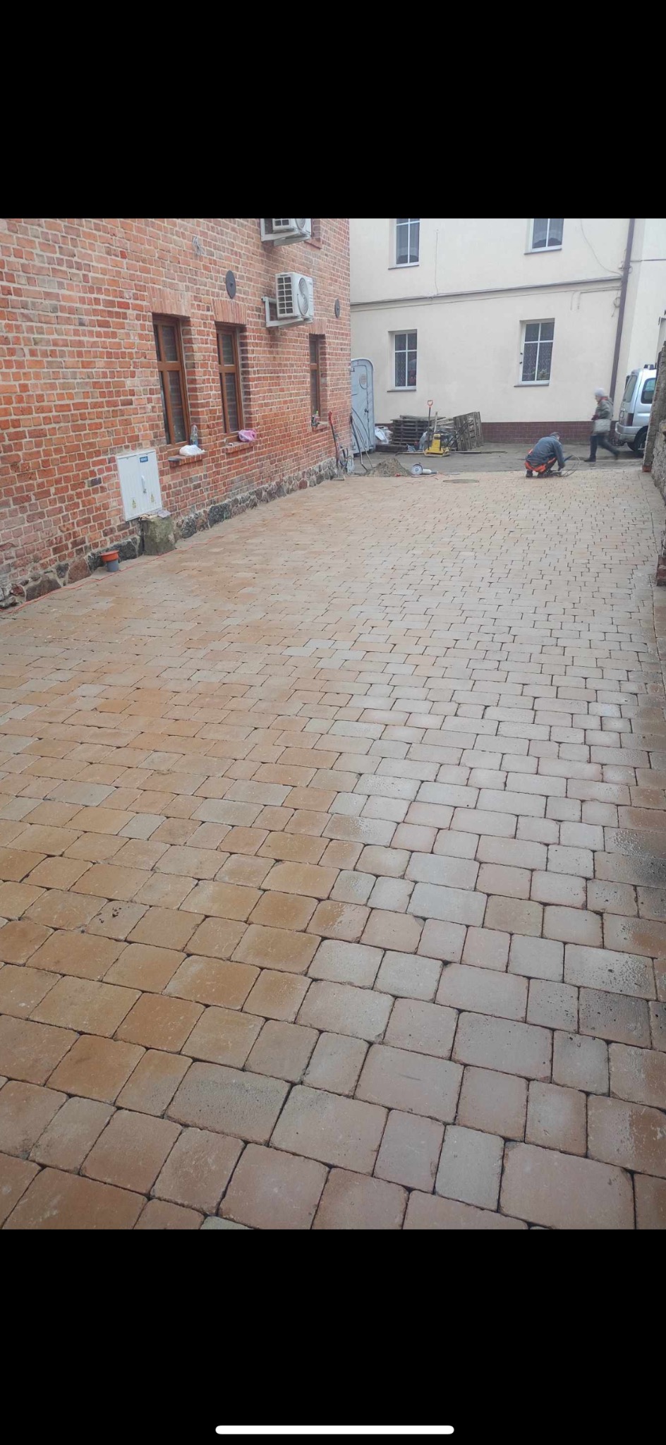 Dziedziniec wyłożony brązową kostką brukową, widoczni pracownicy podczas prac wykończeniowych, budynki z cegły i otynkowane w tle.