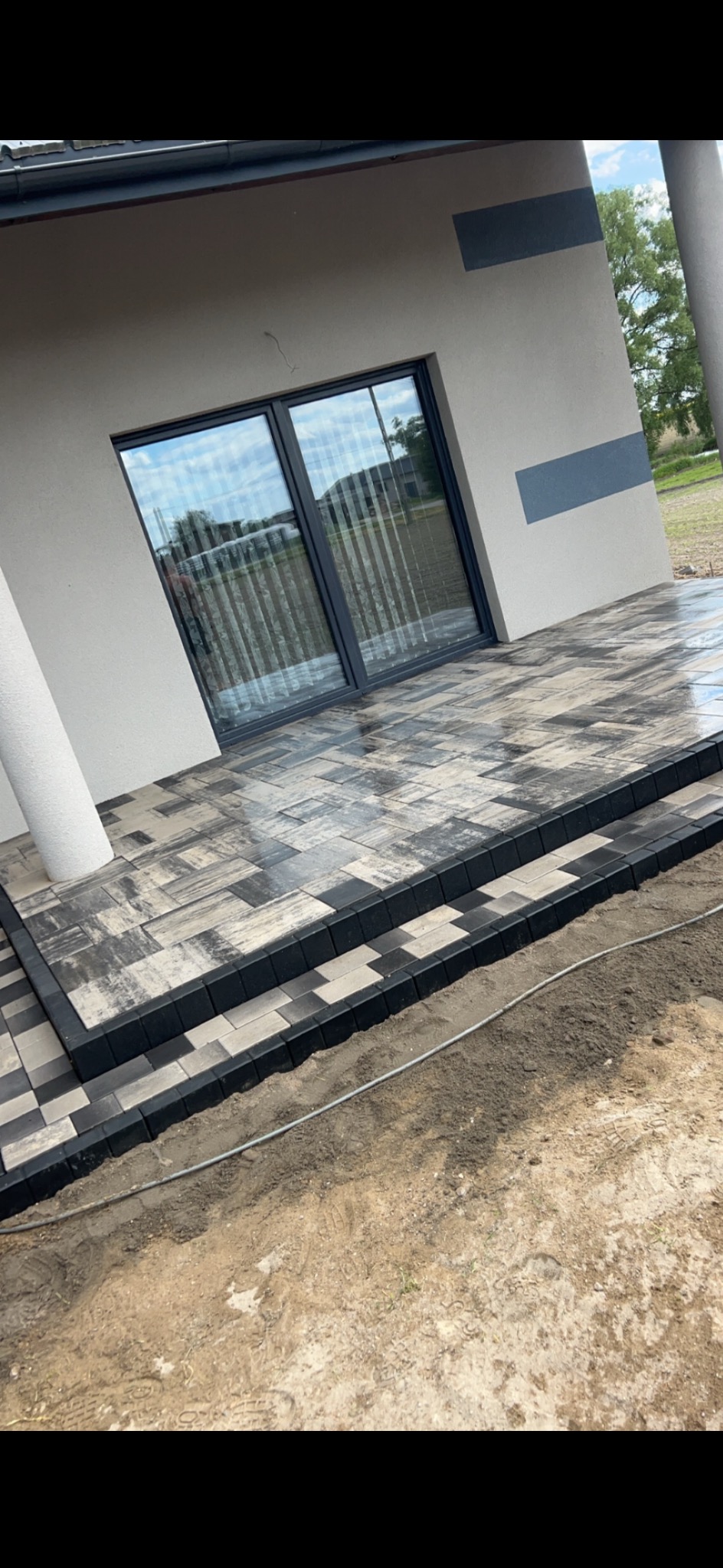 Nowo ułożony taras z kostki brukowej w odcieniach szarości, z widocznymi dwoma stopniami i obrzeżami z ciemnej kostki, przylegający do ściany budynku z dużym oknem tarasowym.