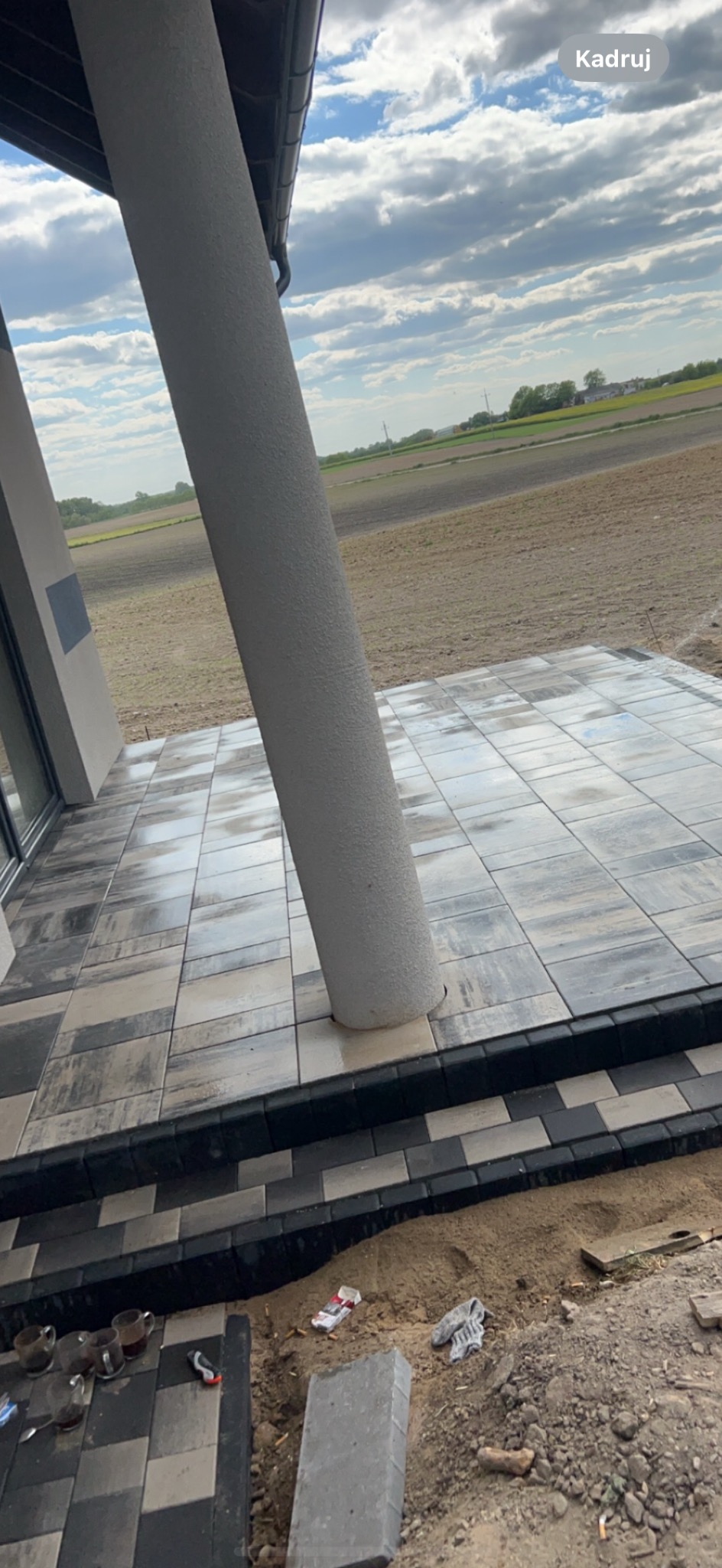 Ułożona, wilgotna kostka brukowa w odcieniach szarości na tarasie z betonową kolumną, widoczne niedoskonałości i ślady budowy: niedopite napoje, niedopałki papierosów i brudne rękawice robocze.