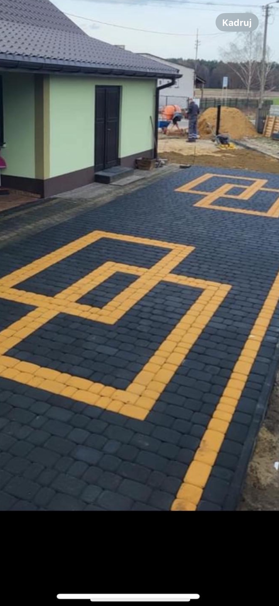Ułożona nawierzchnia z ciemnoszarej kostki brukowej z geometrycznym wzorem z jasnej kostki, widoczny budynek gospodarczy i pracownicy w tle.