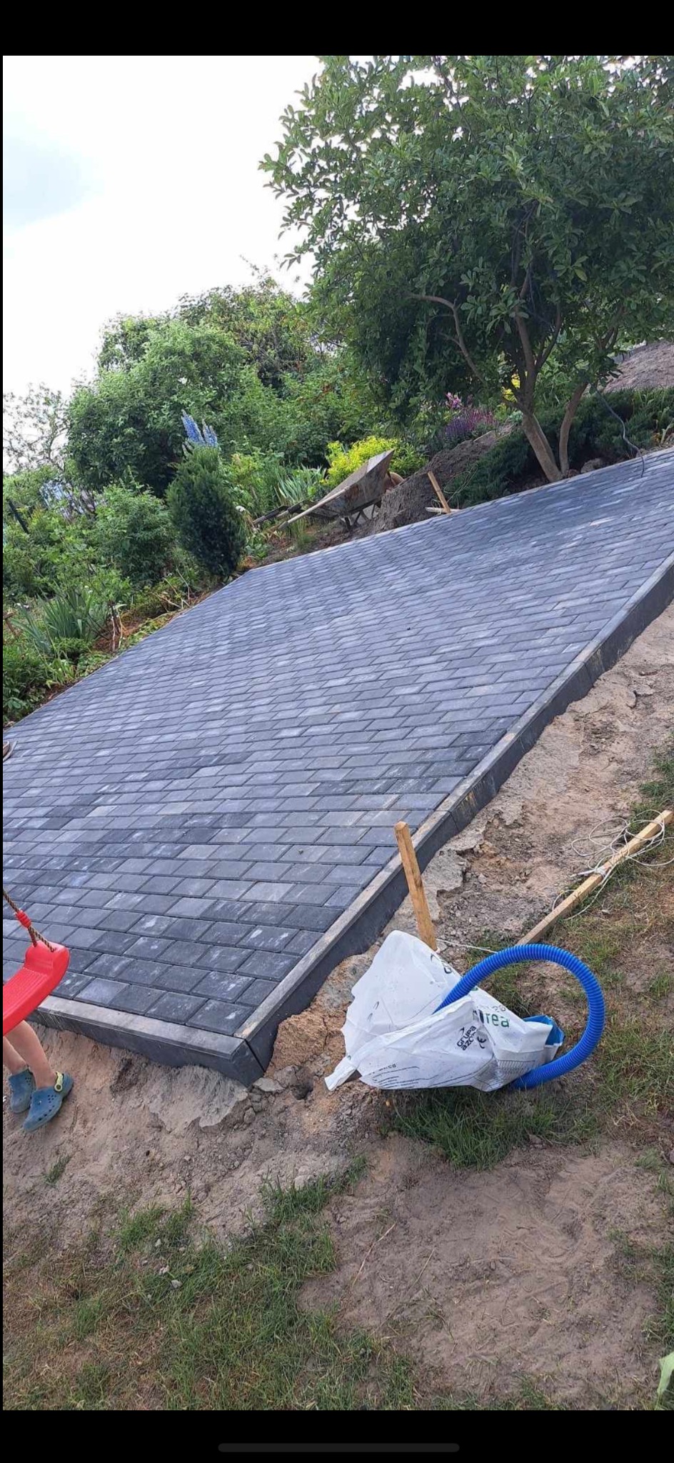 Powierzchnia wyłożona szarą kostką brukową na nierównym terenie, widoczny fragment huśtawki i niebieski wąż, w tle zieleń drzew i krzewów.