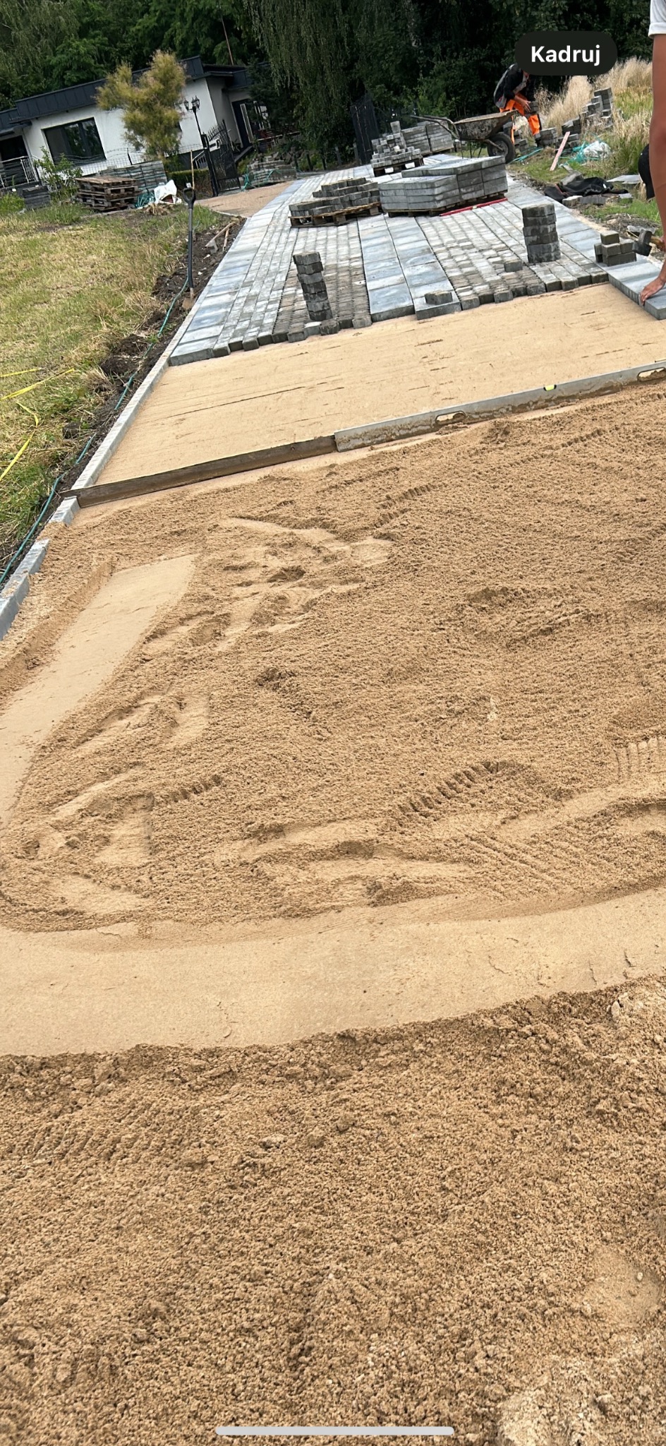 Przygotowany teren pod ścieżkę z kostki brukowej, wyrównany piasek z widocznymi śladami opon, kostka ułożona częściowo w oddali, osoba w pomarańczowych spodniach w tle.