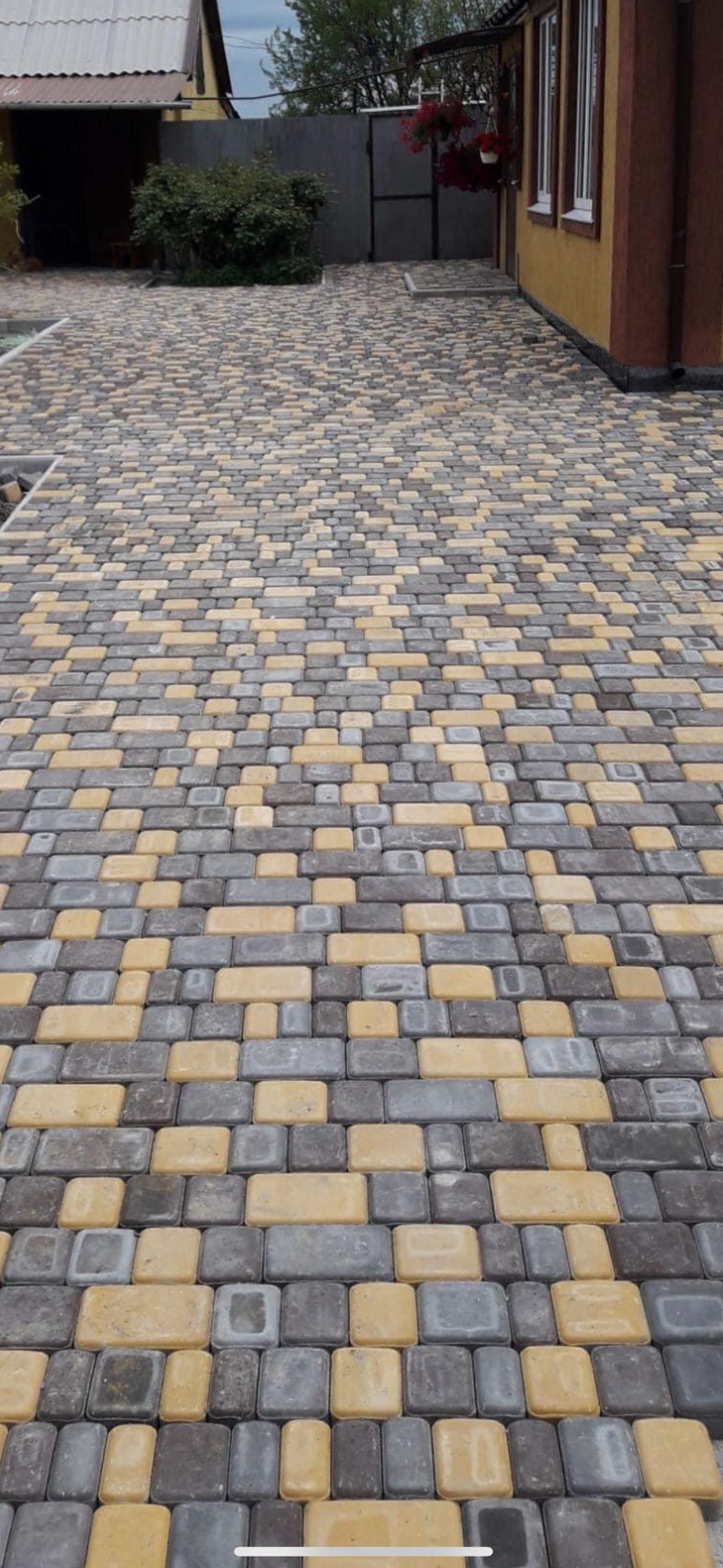Ułożona nawierzchnia z kostki brukowej w odcieniach szarości i żółci, prowadząca do domu z czerwonymi kwiatami w donicach, widoczne złączenia kostek.