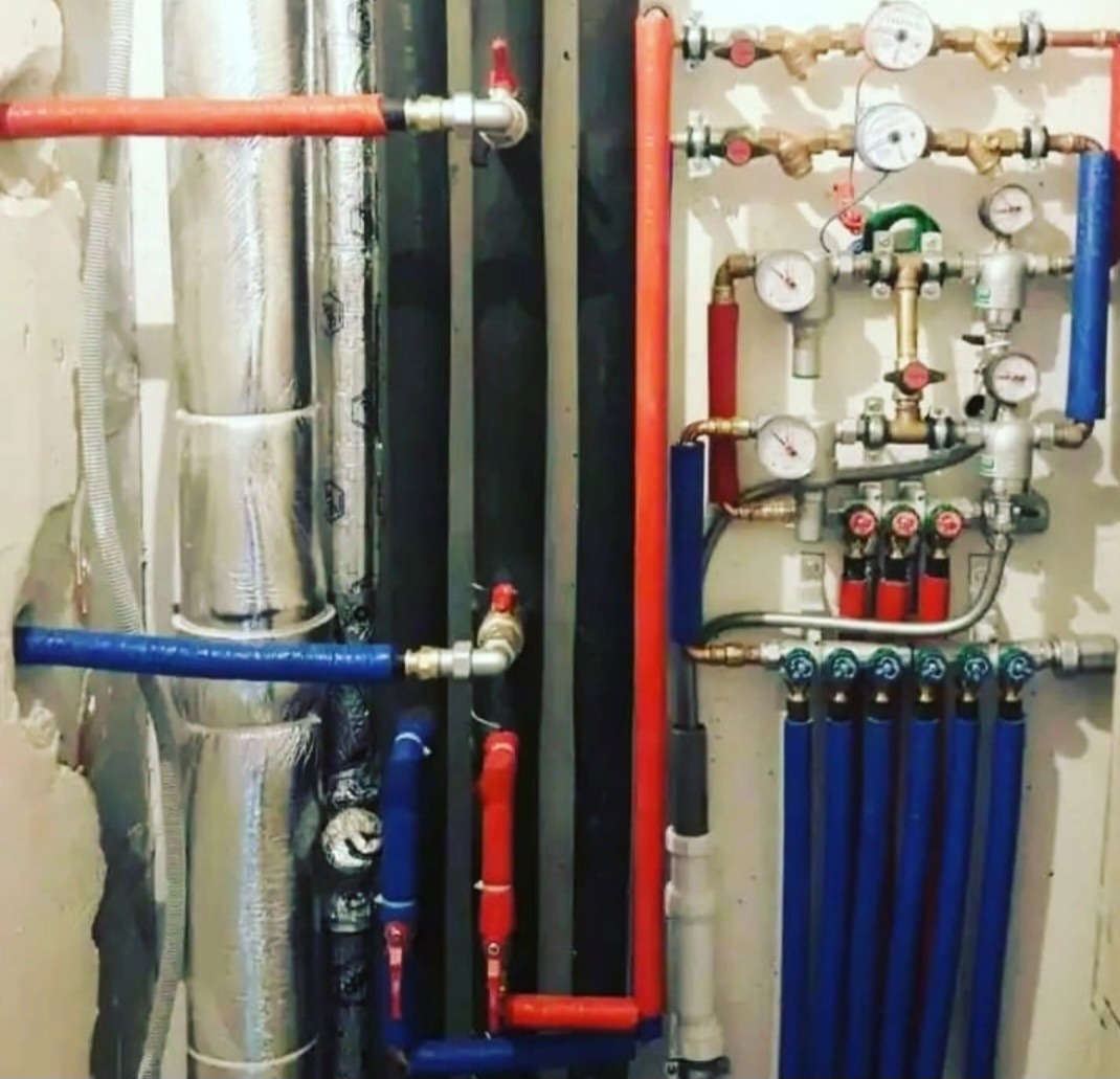 Rozbudowana instalacja wodna z rurami w kolorach czerwonym i niebieskim, manometrami i zaworami, widoczne izolowane rury i elementy montażowe.