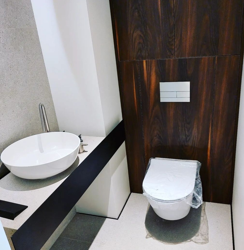 Nowoczesna łazienka z owalną białą umywalką nablatową, baterią chromowaną, toaletą w folii ochronnej i zabudową z ciemnego drewna.