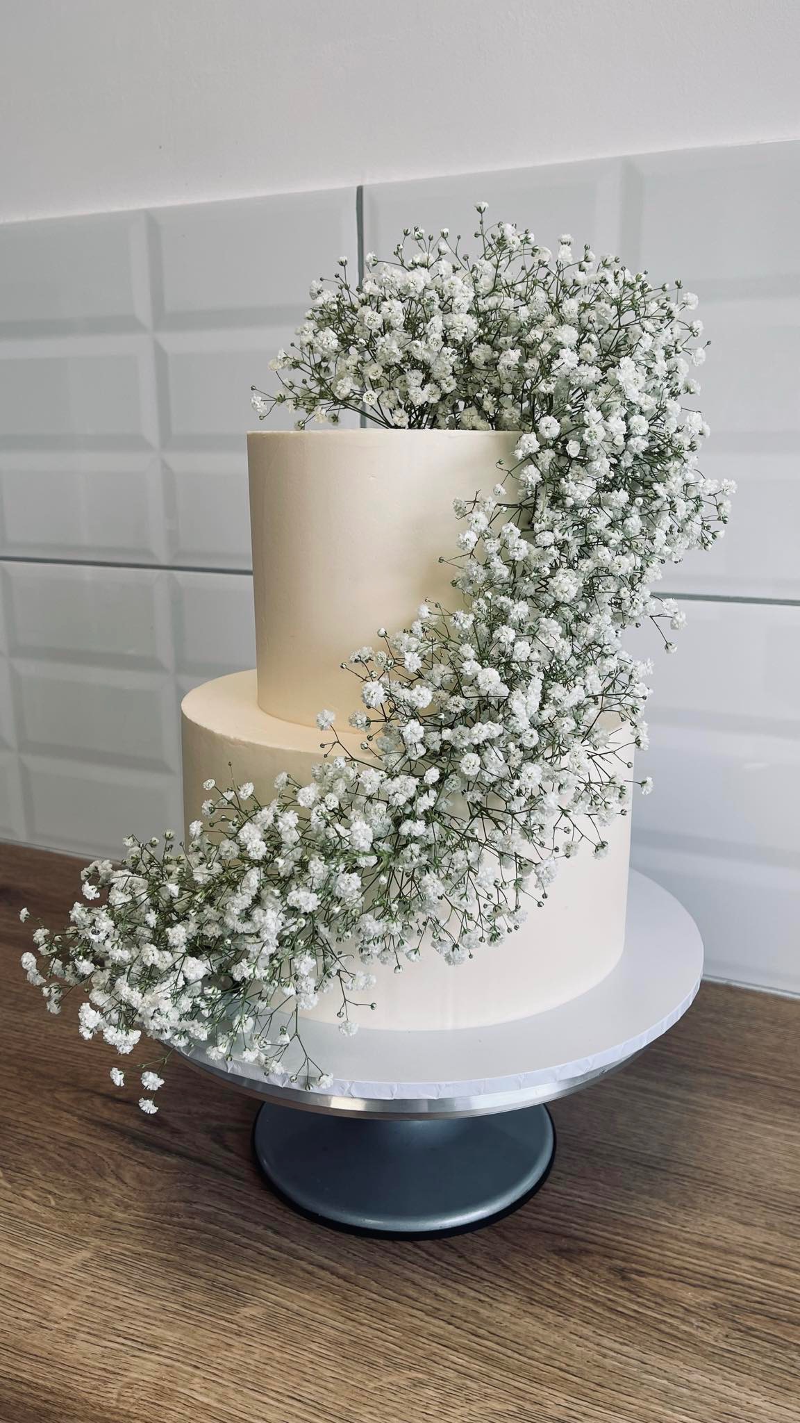 Dwupiętrowy tort weselny w kremowym kolorze, ozdobiony kaskadą białych gipsówek, stojący na srebrnym stojaku obrotowym.