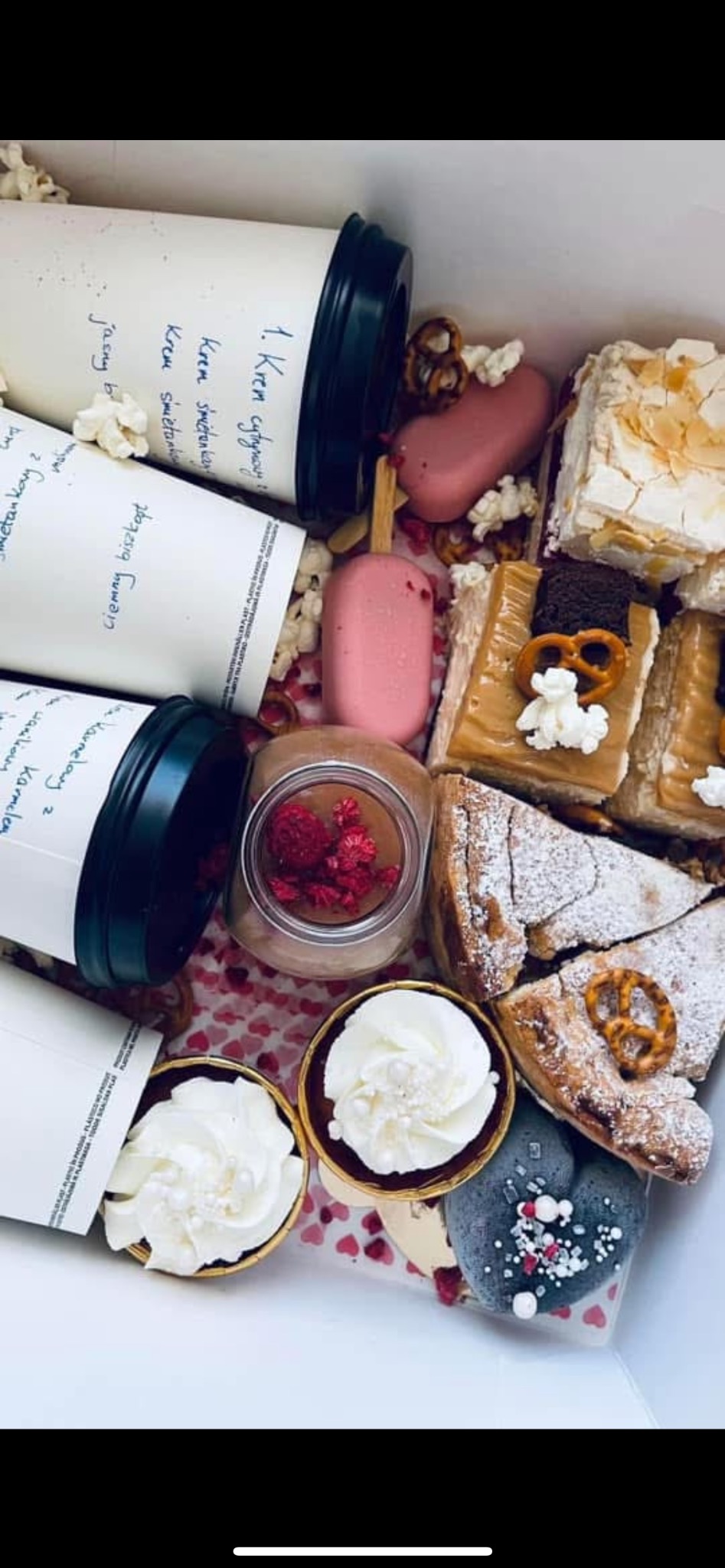 Zestaw słodkości w pudełku: ciasta, desery w słoiczkach z malinami, babeczki z kremem, lizaki w kształcie serca, precelki i popcorn, obok stoją kubki z wypisanymi składnikami.