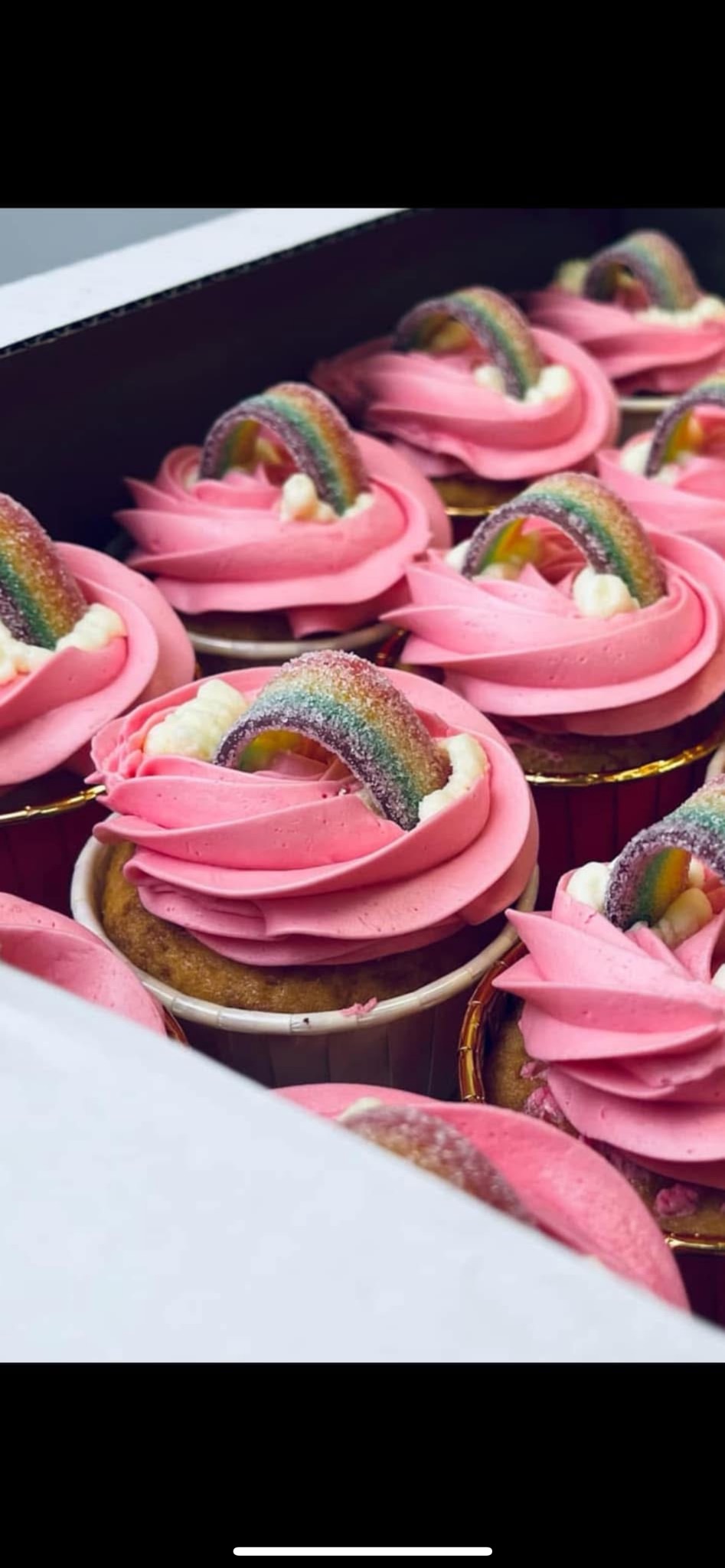 Karton z rzędem babeczek z różowym kremem i żelkowymi tęczami, widoczne złote papilotki.