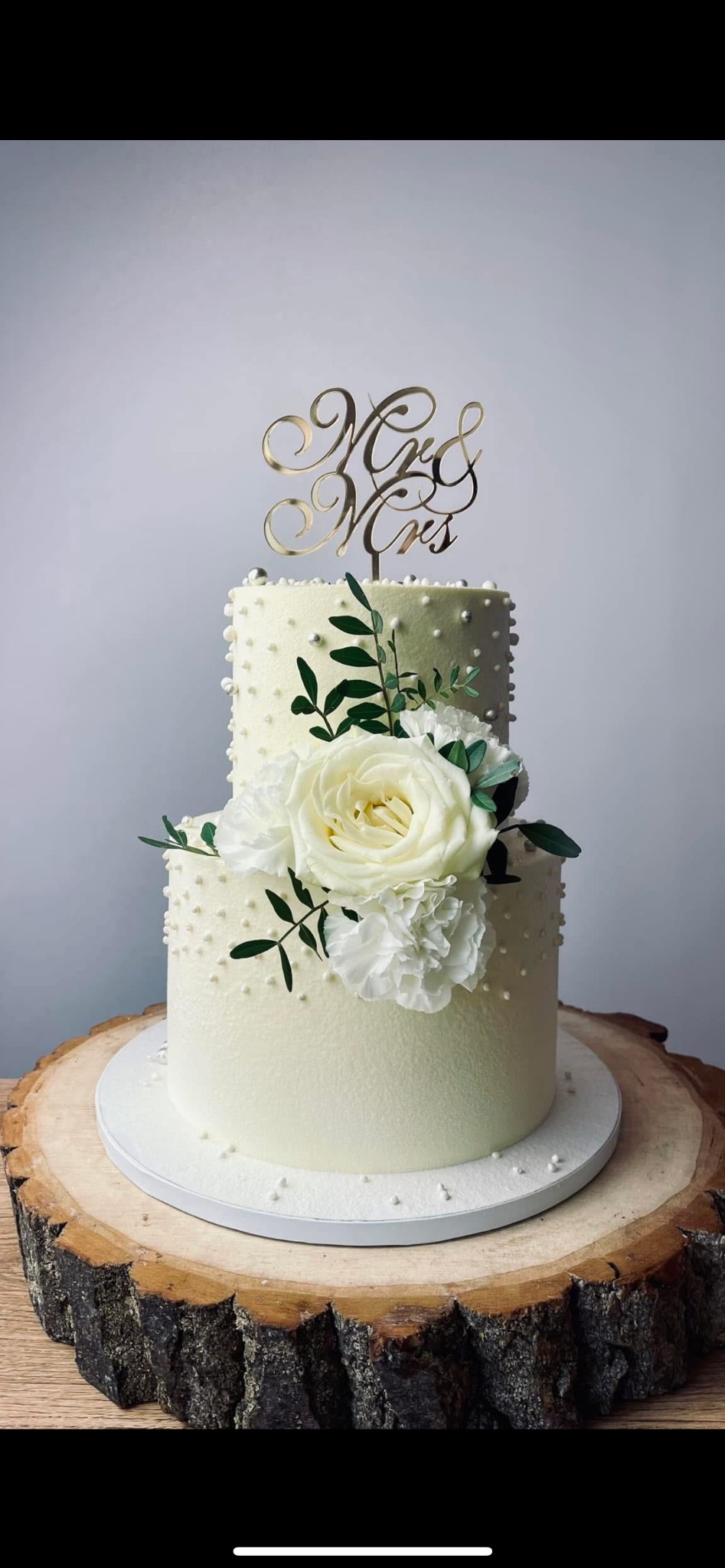 Dwupoziomowy tort weselny w białej polewie, zdobiony białymi różami i zielonymi listkami, z topperem 'Mr & Mrs', umieszczony na drewnianym plastrze pnia.