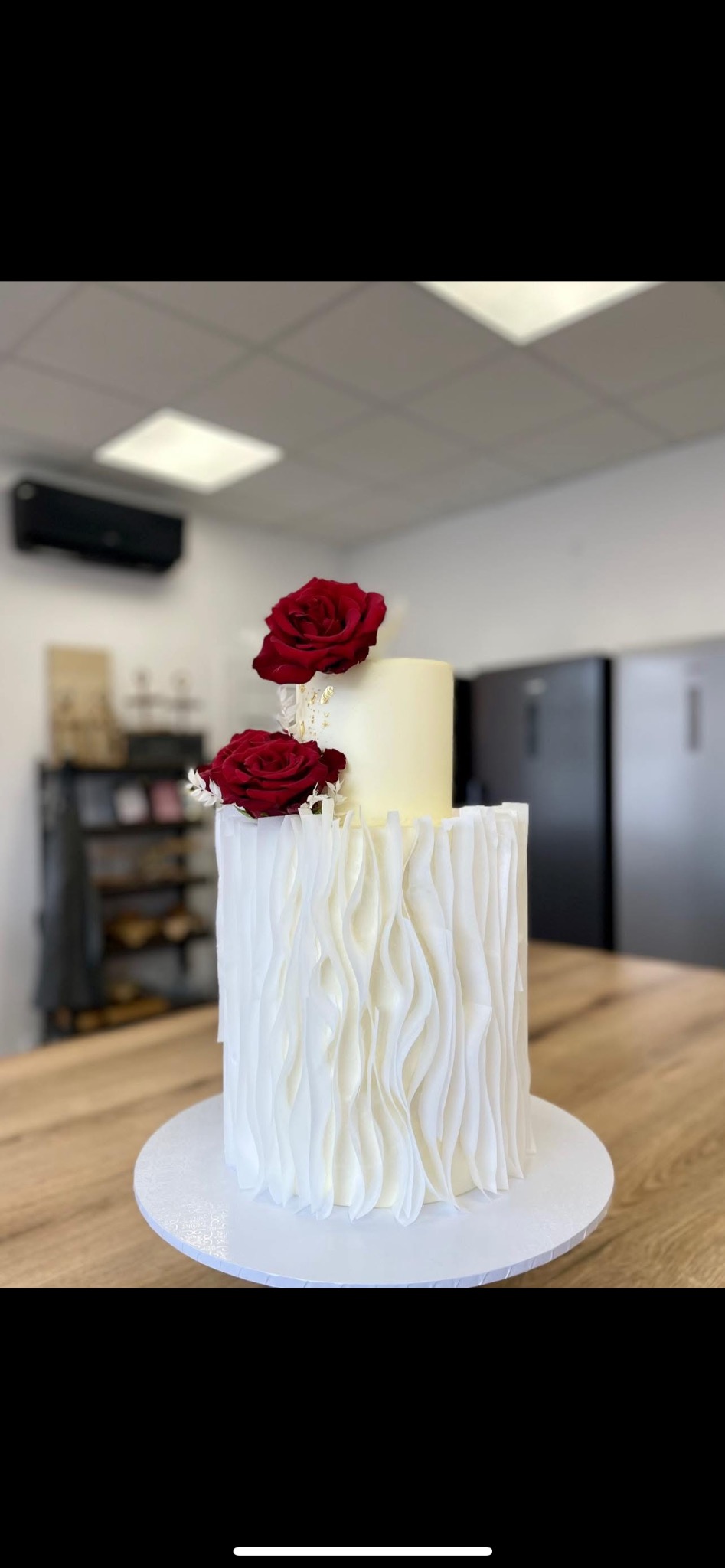 Dwupiętrowy tort weselny w odcieniach bieli, z falistą dekoracją i dwiema czerwonymi różami na górze, ustawiony na drewnianym stole w jasnym pomieszczeniu.