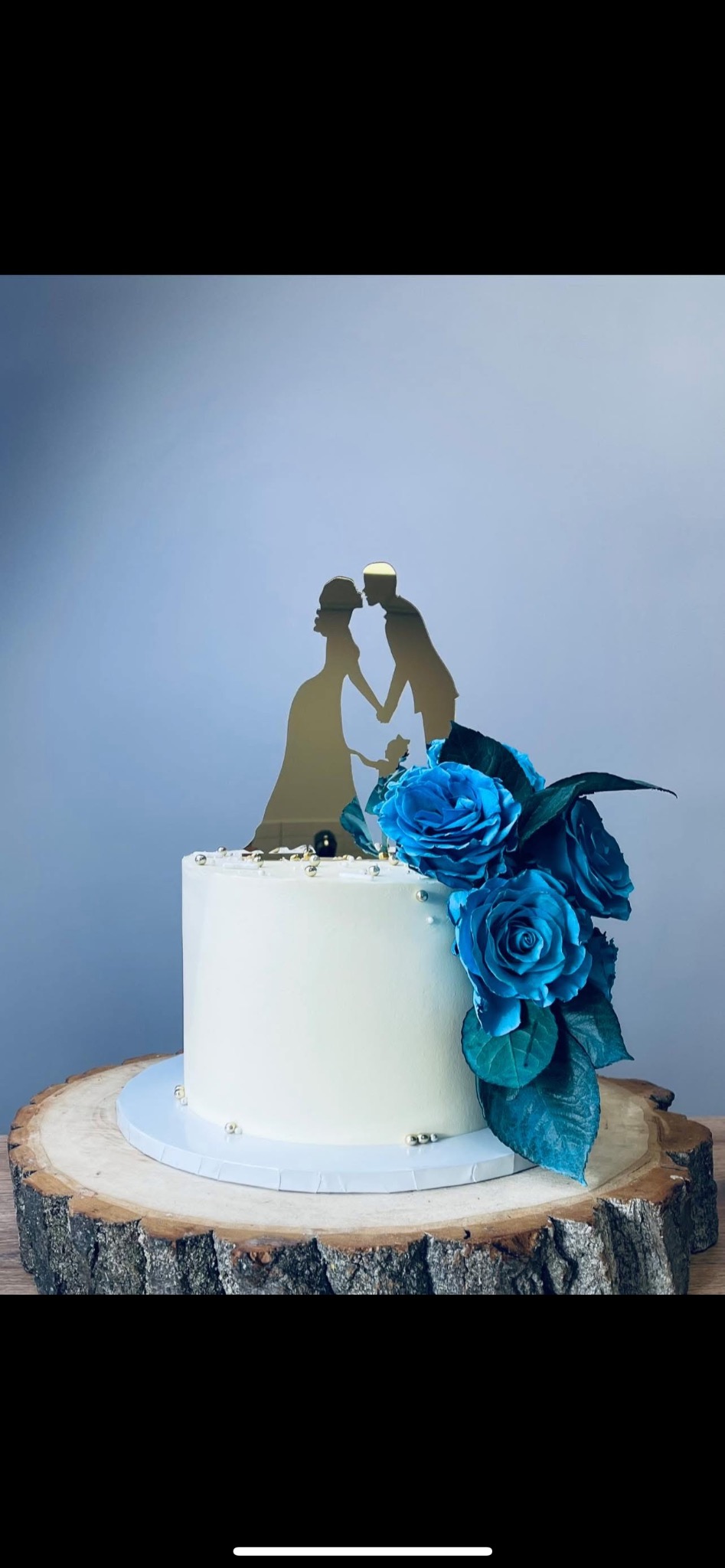 Tort weselny z białym kremem, dekorowany niebieskimi różami i złotą sylwetką pary młodej z dzieckiem, umieszczony na drewnianym plastrze.