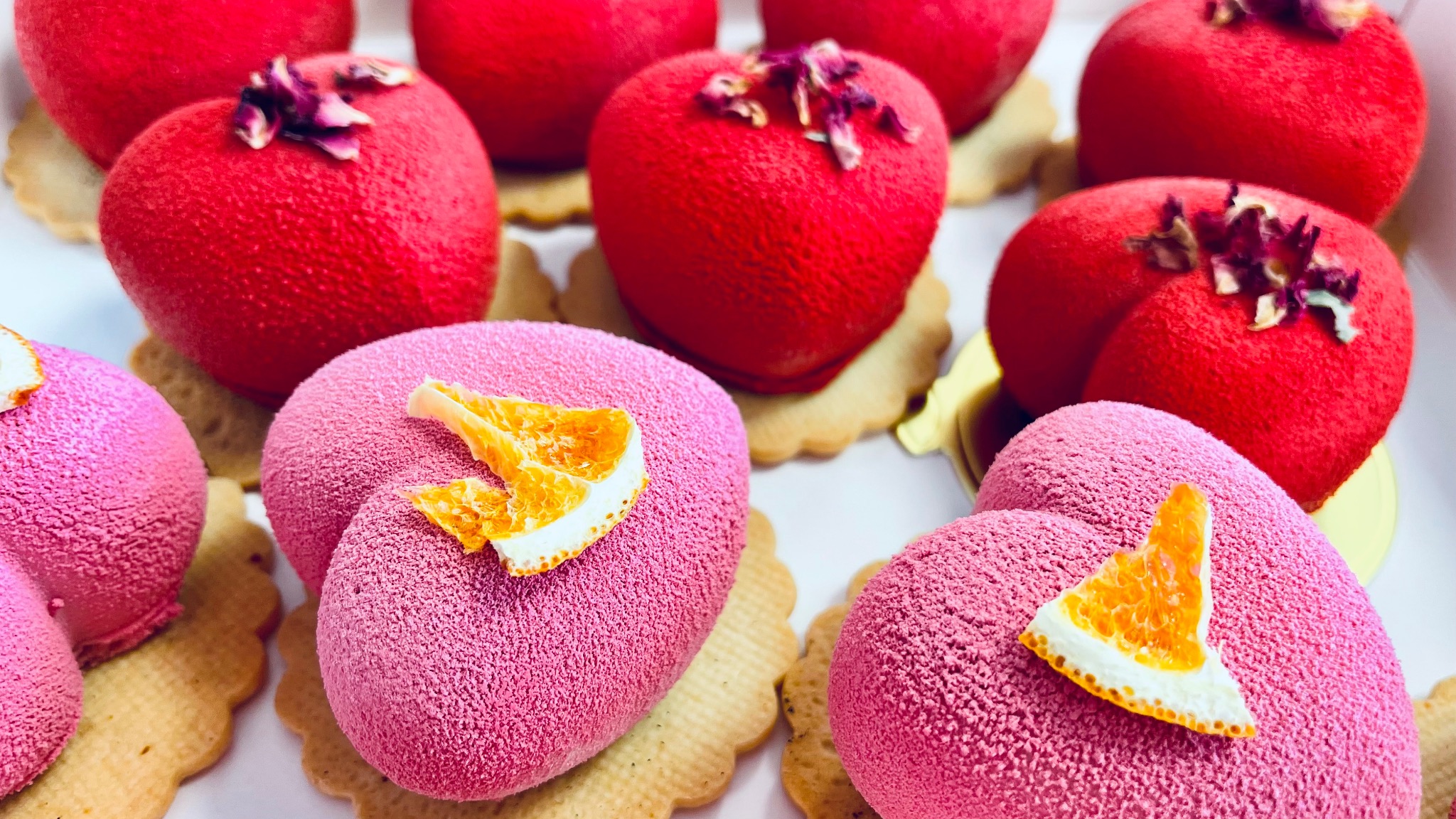 Różowe i czerwone deserowe serca z aksamitną teksturą, na kruchych spodach, udekorowane kandyzowaną skórką pomarańczy i suszonymi płatkami róż.